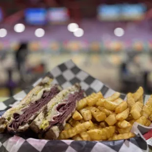 Hot pastrami on rye with a side of perfectly seasoned fries, with a blurry view of the bowling alley in the background