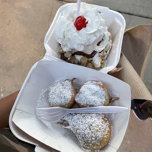 a hand holding a container of powdered sugared donuts with a cherry on top