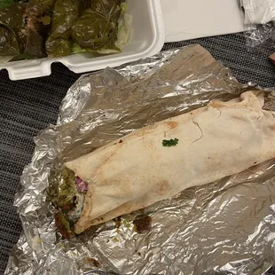 a burrito wrapped in tin foil next to a container of stuffed grape leaves