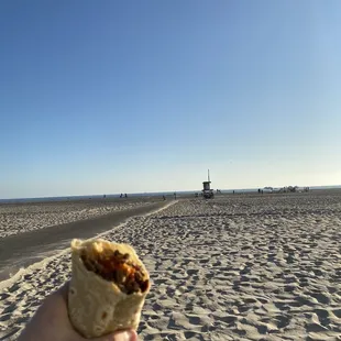 a hand holding a burrito on the beach