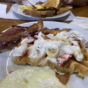 Waffle topped with bananas and strawberries eggs and bacon