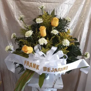 White and yellow sympathy spray in a paper mache basket. Roses, carnations, solidago, dracaena, palm leaf, leather leaf, white bow, banner