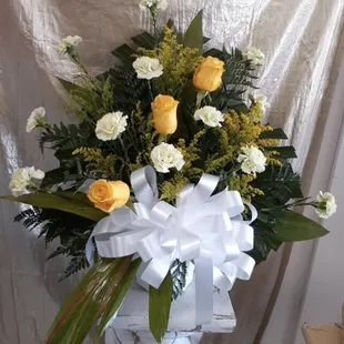 White and yellow sympathy spray in a paper mache basket. Roses, carnations, solidago, dracaena, palm leaf, leather leaf, white bow
