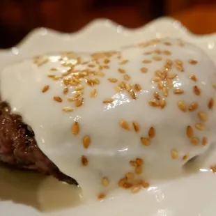 a hamburger covered in white sauce and sprinkled with sesame seeds