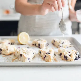 a woman sprinkles icing on a tray of blueberry scones