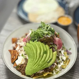 Chilaquiles: chips w/green &amp; red salsa, crema, queso fresco, red onion, avocado, radish &amp; cilantro w/beans &amp; 2 eggs | $18 + Short Rib $6