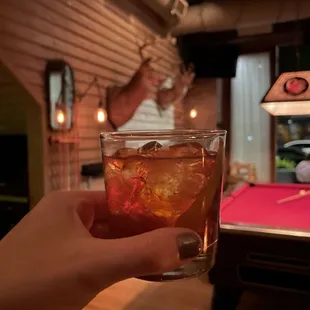 a person holding a drink in front of a pool table