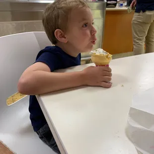 a young boy sitting at a table eating an ice cream cone