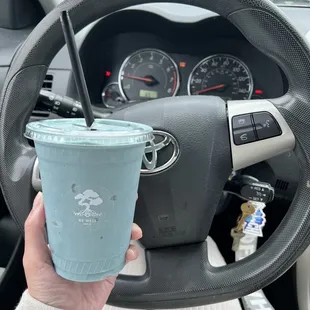 a hand holding a blue drink in front of a steering wheel