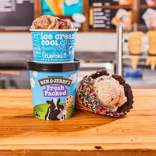 Waffle cone, cup and fresh packed pint in front of Ben &amp; Jerry&apos;s ice cream shop menu board.