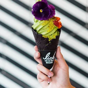 Matcha soft serve in a midnight vanilla cone topped with strawberries , pistachio, edible flower , &amp; dusted with matcha  powder.