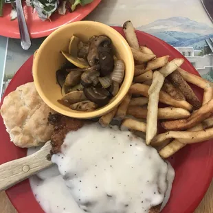 Chicken fried chicken with a side of sautéed mushrooms and onions alongside a biscuit and French fries.