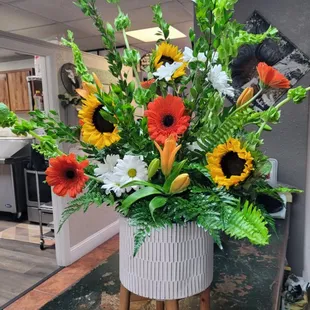 3 legged plant stand. Floral design of Bells of Ireland, orange gerbera daisy's and roses, sunflowers, lily's and assorted greens.