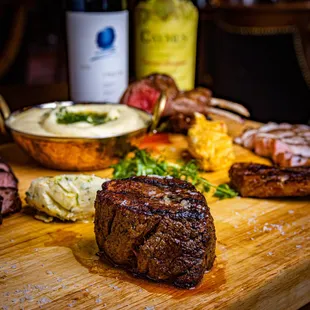a wooden cutting board with steak, potatoes, and a bottle of wine