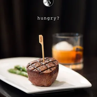 a steak on a white plate with a glass of whiskey in the background