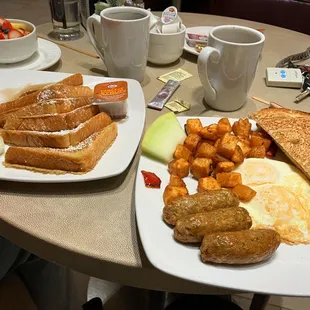 On the left, fresh fruit and French toast. Plate on the right is two eggs, fried seasoned potatoes, sourdough toast and 3 chicken sausages.