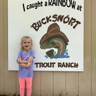 a little girl standing in front of a sign that says caught a rainbow at bucksnort trout ranch