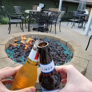 two people holding beer bottles in front of a fire pit
