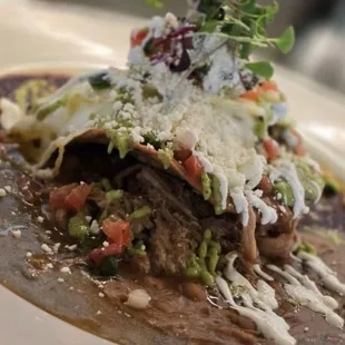 Two crunchy corn tortillas on top, green chili and adobo sauce and beans, pork Carnitas eggs, avocado, salsa, and pico de gallo