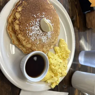 Pancakes and scrambled eggs with a side of maple syrup.