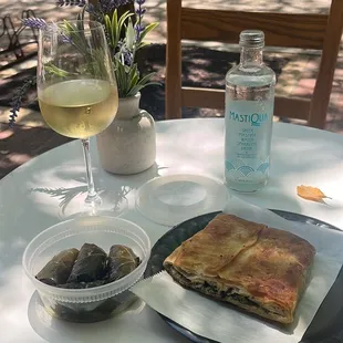 (Left to right) Glass of white wine, Mastiqua sparkling water, vegan dolmades (grape leaves with rice and herbs inside), spinach pie