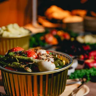 A close-up of a golden bowl of roasted onions and tomatoes, with a spread of fruits and a bowl of mashed potatoes in the background.