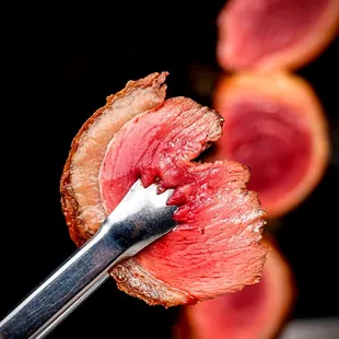 A perfectly cooked slice of Picanha, held by a pair of tongs, ready to be enjoyed.