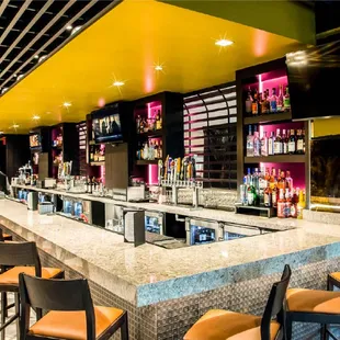 The image shows a bar with a friendly bartender standing behind the counter. The bar has lots of different alcohol bottles ne...