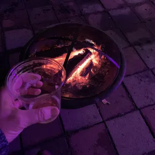 a person holding a glass of beer in front of a fire pit