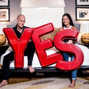 a man and a woman sitting on a bed holding a yes sign