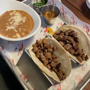 Carne asada street tacos with a side of refried beans. Included in the small cups is a side of cilantro with diced onions and taco sauce.