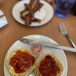 English Muffin with butter and homemade strawberry jam with a side of bacon in the background.