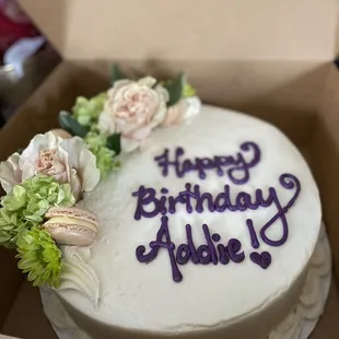 White birthday cake with purple &quot;Happy Birthday, Addie!&quot; icing message, live pink and green flower, macaron, and icing decorations.