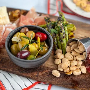 a wooden cutting board with a bowl of olives, nuts, asparagus, and prosciutto