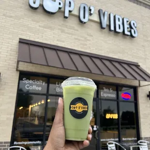  a hand holding a cup of green tea outside a coffee shop