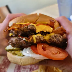 a hand holding a hamburger with bacon, cheese, tomato, and pickles