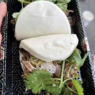 a hand holding a black plastic container with a white bun and green garnish on top