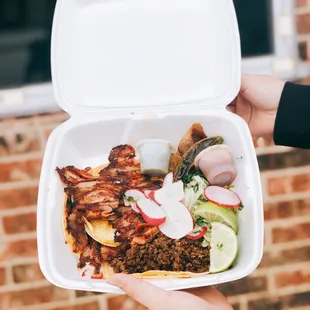 a person holding a takeout box with a taco and salad