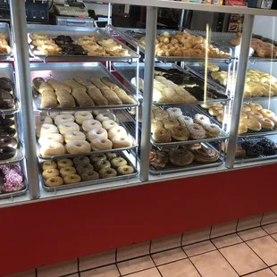 Picture of donuts listed in rows on trays. Nicely cleaned, organized and well lit in the cases.