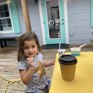 a little girl sitting at a table with a cup of coffee