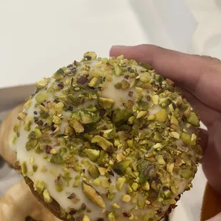 a hand holding a donut with white icing and pistachios