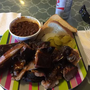 Beef brisket, chicken and ribs dinner plate with pickles, onions and beans.
