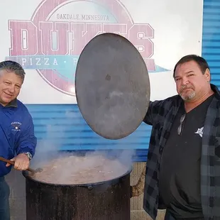 two men standing in front of a large pot of food