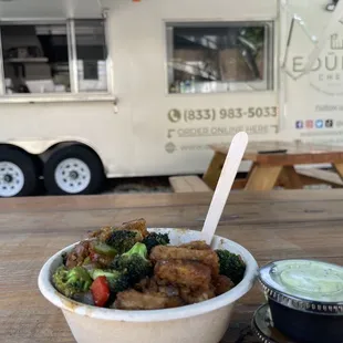 a bowl of broccoli and meat in front of a food truck