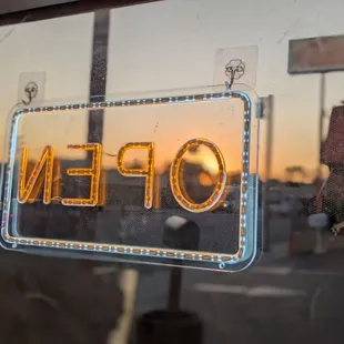 "Open" sign facing East, and it is lit up. The sunset is seen in the background through the opening of the letter 'O'.