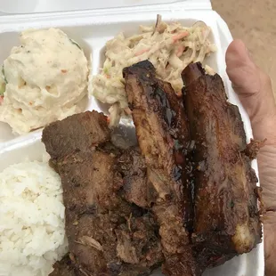 a hand holding a styrofoam container with ribs, rice, coleslaw and coleslaw