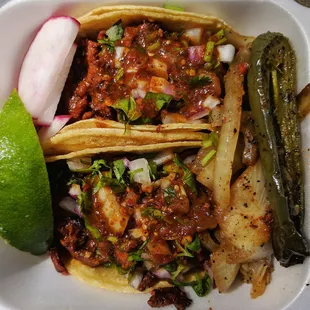 Two pork tacos with cilantro, onion, and hot sauce. Radishes, piece of lime, grilled onions and half jalapeno on the side.