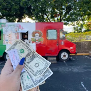 a hand holding money in front of a food truck