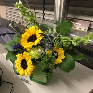 Flower arrangement sitting on a desk.  Primarily sunflowers and greenery, in a cute square glass vase.