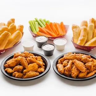 two plates of chicken wings, carrots, celery sticks, and dips